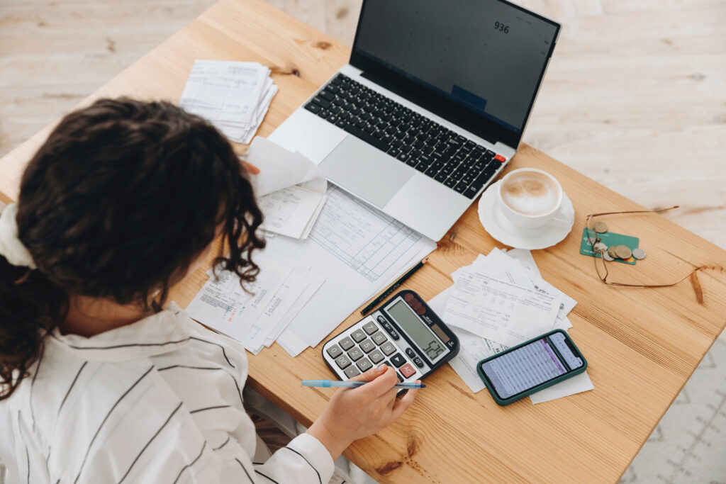 Top view of a woman researching how much does small business bookkeeping cost with bills, calculators and a laptop in front of her